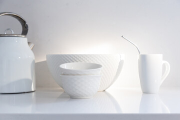 Selective focus view of white ceramic bowls, coffee cup with metal straw and kettle set on quartz countertop in rays of sunlight