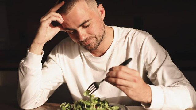  Man wear white long sleeves sitting at table, looking sad and bored with diet not wanting to eat salad. Modern kitchen interior. Healthy nutrition, dieting, eating disorder.