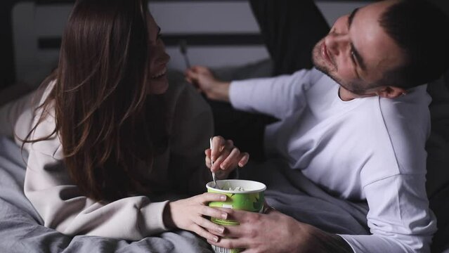 Cheerful couple eating pistachio ice cream lying in the bed at home. Woman feeding her boyfriend with spoon. Dating. Couple have fun at home. Love concept.