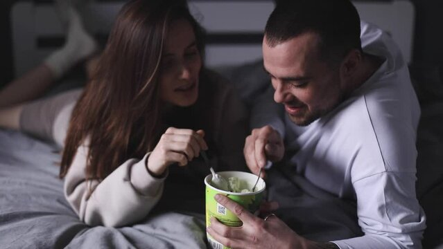 Cheerful couple eating pistachio ice cream lying in the bed at home and talking. Woman feeding her boyfriend with spoon. Dating. Couple have fun at home. Love concept.