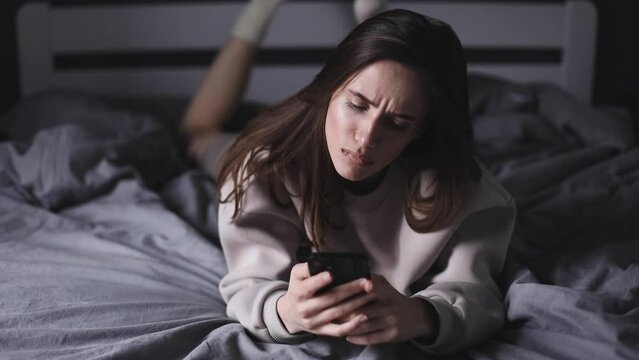 Sad woman lying in a bed in bedroom and uses smartphone. Girl chatting use her mobile phone, she look nervous and unhappy.