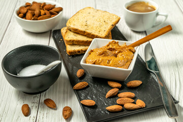 Raw Organic Almond Butter in a bowl on table with wooden spoon and almonds scattered on the table, toast bread and coffee