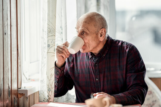 Grandfather drinking coffee or tea from white cup and looking through window. Lonely retired man have breakfast.