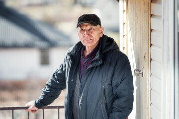 Older farmer with pensive look stands on porch of cottage house outside. Ordinary rustic senior man...