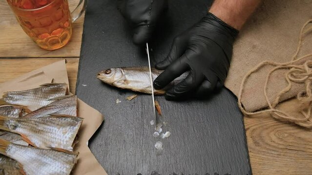 A cook in black latex gloves, cuts on a black kitchen board dried fish roach with caviar in half. Shows the cut halves to the camera and lays next to another chopped fish. The table is served with a g
