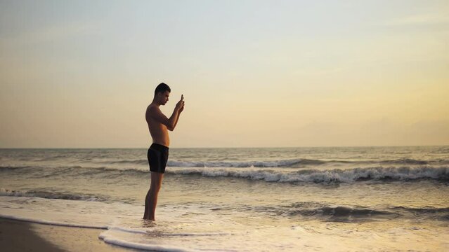 Handsome shirtless young man using smartphone to take photo of the sunset beach, standing on the sea shore