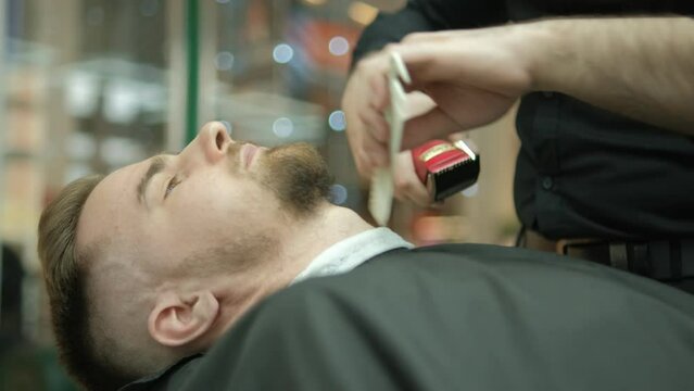 Trendy bearded barber cuts bearded man's hair with a clipper in barbershop. Men's hairstyling and hair cutting in salon. Grooming the hair with trimmer. 4K