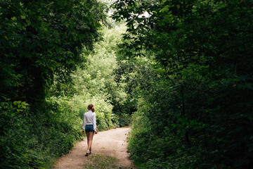 Obraz premium A young slender schoolgirl in denim shorts and a white shirt walks along a forest path into the distance, under an arch of trees.