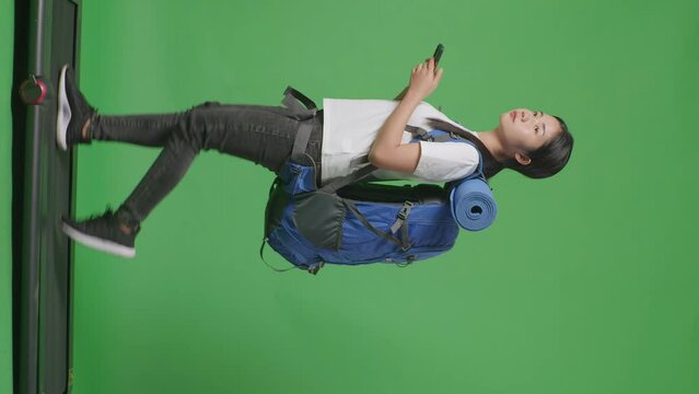Full Body Side View Of Asian Female Hiker With Mountaineering Backpack Looking At The Map While Walking On A Treadmill On Green Screen Background In The Studio
