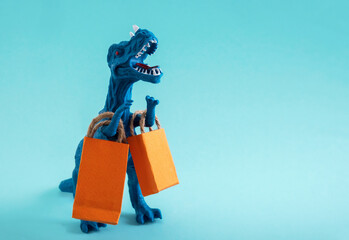 Cute blue dinosaur with orange shopping bags on a blue background. Cute humor shopping concept. Copy space. - obrazy, fototapety, plakaty
