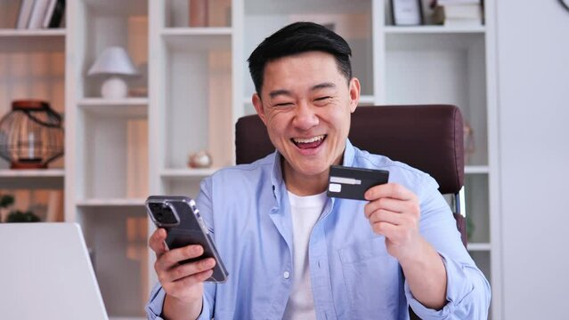 Asian freelancer celebrates success holding credit card phone in office making online payments through bank card on smartphone. Sports betting aspect highlights financial rewards of smart investments.