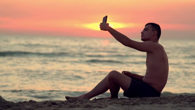 Young handsome man sitting on the sandy tropical beach taking a selfie while on vacation relaxing and enjoying the sunset view