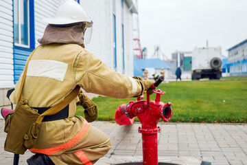 Fire rescuer in helmet and protective clothing sits next to fire hydrant outside on street. View...