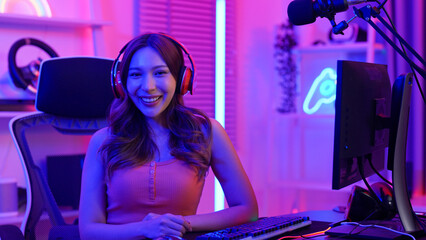 Asian young adult woman is looking at camera with smile face and introducing herself before playing a game with live streaming. A female gamer is wearing headphones and enjoying playing online game.