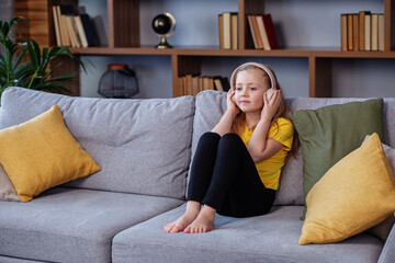 Obraz premium Happy child listens to music or audiobook at home on couch. Little school kid wearing headphones
