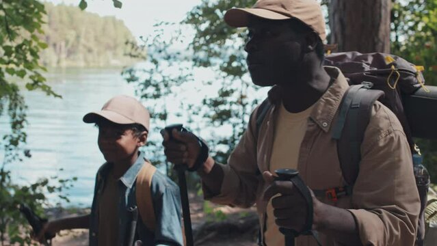 Tracking shot of active African American senior man and his grandson wearing backpacks hiking along lake shore using trekking poles