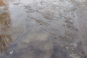 The top down, close up view of a giant pot hole that is filled with water from the melting snow bank in front of it.
