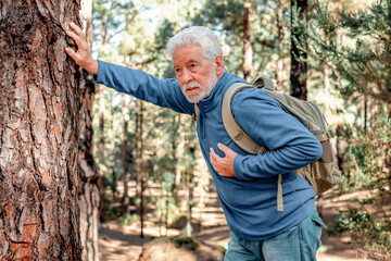 Old Senior Bearded Man in a Trekking Day in the Mountain Woods Stops in Pain in his Heart and Leans Against Tree Trunk to Catch his Breath