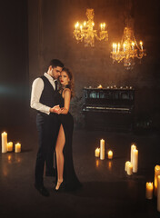 sexy couple blonde woman holding hand of handsome man slow dance in dark gothic room piano, candles are burning, candlelight holiday party. Guy hugs girl. Black evening dress art beauty face red lips 
