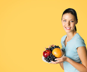 Portrait image of young happy smiling attractive woman in blue casual smart clothing, with plate of fruits, isolated over orange yellow background, with copy space area for text.