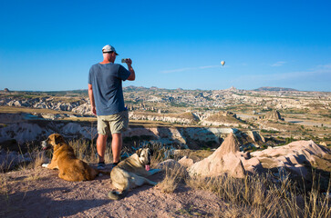 Travel to Cappadocia, Anatolia region, Turkey. A male tourist stands on a hill overlooking the Cappadocian landscape, two large dogs lie on the ground