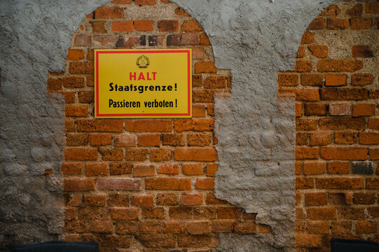 Eine historische Ziegelmauer mit einem gelben Warnschild, auf dem &ldquo;Halt! Staatsgrenze! Passieren verboten!&rdquo; steht, als Hinweis auf vergangene Grenzmarkierungen
