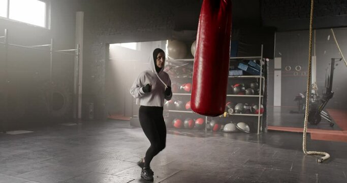 Zooming out beautiful Caucasian female wearing hood and sports outfit with boxing bandages, training in fitness club. Charming woman punching and beating boxing bag. Concept of sport routine.