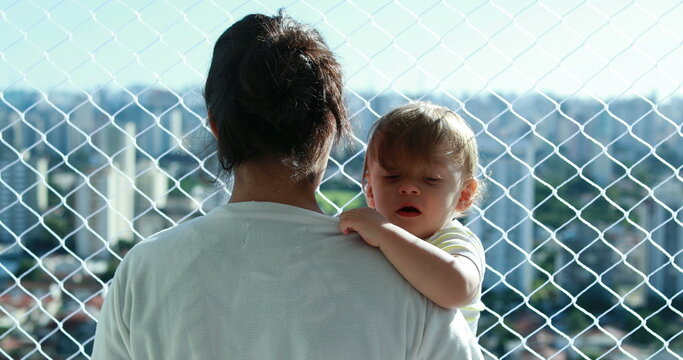 Mother holding baby overlooking city at apartment balcony with safety net window