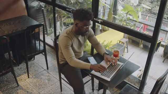High angle shot of young black man in casualwear and glasses working on laptop while doing freelance job at table in modern cafe