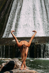 Naklejka na ściany i meble A slender tanned girl with a sports figure is doing yoga exercises against the backdrop of a picturesque waterfall in the summer. She is wearing black shorts and a top.