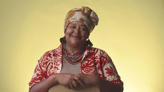 Portrait of cheerful mature African American woman in bright turban and Hawaiian print shirt posing for camera on yellow background