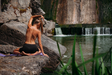 Fototapeta na wymiar A beautiful tanned girl with a sports figure is doing yoga exercises against the backdrop of a picturesque waterfall in the summer. Sits in the lotus position. She is wearing black shorts and a top.