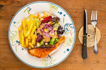 Top view of delicious homemade salmon steak with french fried and caesar salad in white ceramic plate with table knife and metal fork on bamboo mat on wooden table.