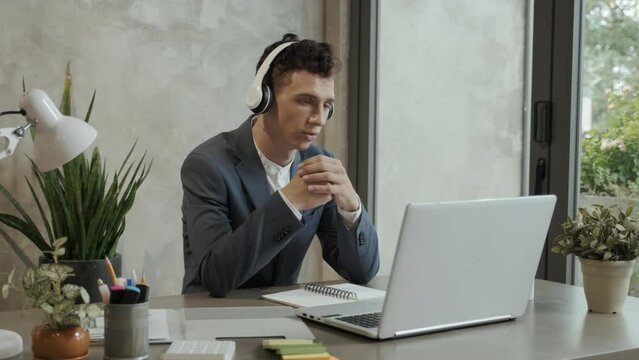 Young elegant businessman in headphones sitting by workplace in front of laptop, looking at screen and showing five fingers while describing business points