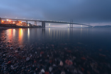 Two bridges covered in thick fog at sunset in winter, and colourful stones in the foreground. Forth...