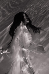 Underwater shoot of beautiful woman in white flying transparent dress relaxing in water in sunbeams.