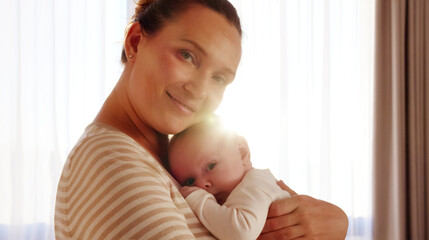 Young happy  mother  holds her newborn baby, window with sunset behind on a background. Closeup portrait of a loving mother with a child. Happy mom with her  infant son. Mum loves of her infant child