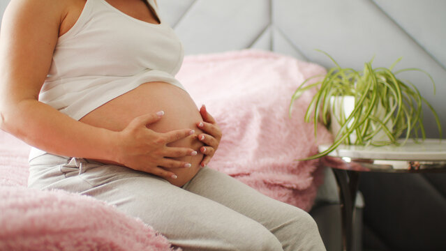 A pregnant woman strokes her stomach with her hand. Pregnant girl sits on the bed and touches her big belly