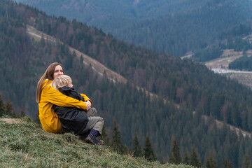 Naklejka na ściany i meble Young woman and child boy sit on hillside. Mother and son rest and sit in an embrace on mountains background.