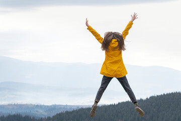 Cheerful girl in bright yellow jacket jumps with hands up to the sky on the mountains background....