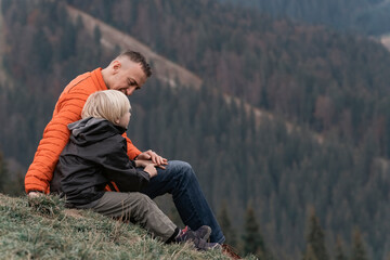 Naklejka na ściany i meble Father and his son sit on slope and look at the mountains. Family trekking with children in forest