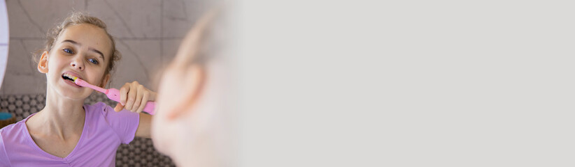 A teenage girl brushes her teeth with an electric toothbrush while standing in the bathroom and looking at her reflection in the mirror. Health and hygiene. Modern oral care. Banner