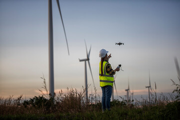 Asian woman in white helmet working with UAV.  at renewable energy farm. Female controlling  UAV on wind turbines outdoors.
