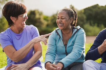 Happy senior female friends having fun together after yoga class at city park - Multiracial elderly women sitting together outdoor