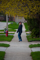 A woman turns to look at the camera while walking on a sidewalk with trees budding in the spring