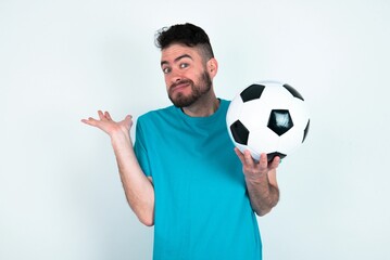 Clueless Young man holding a ball over white background shrugs shoulders with hesitation, faces doubtful situation, spreads palms, Hard decision