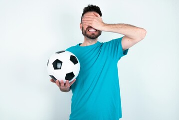 Young man holding a ball over white background covering eyes with hands smiling cheerful and funny. Blind concept.