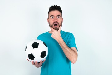 Young man holding a ball over white background Looking fascinated with disbelief, surprise and amazed expression with hands on chin