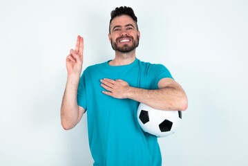 Young man holding a ball over white background smiling swearing with hand on chest and fingers up,...
