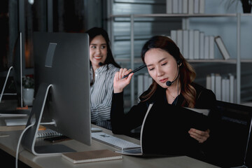 Beautiful Asian woman with headphones work night shift at call center customer care service desk, look at handbook for information to answer question, female with hands-free phone work hard overtime.